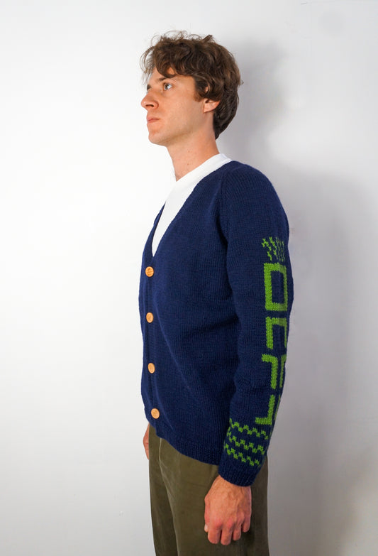 A man wearing a navy blue knit cardigan with green geometric patterns on the sleeve and button accents, standing against a plain light gray wall.