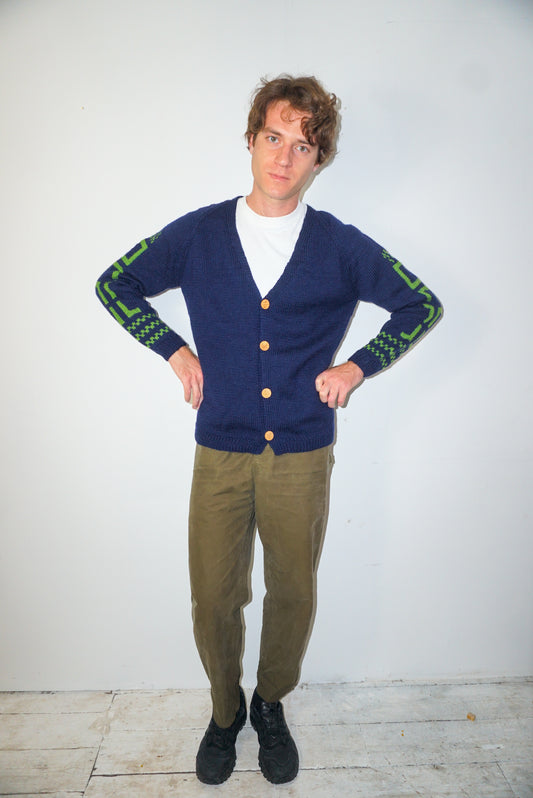 A man modeling a navy blue knit cardigan with green patterned cuffs, worn over a white t-shirt and olive green trousers, standing against a plain white wall.