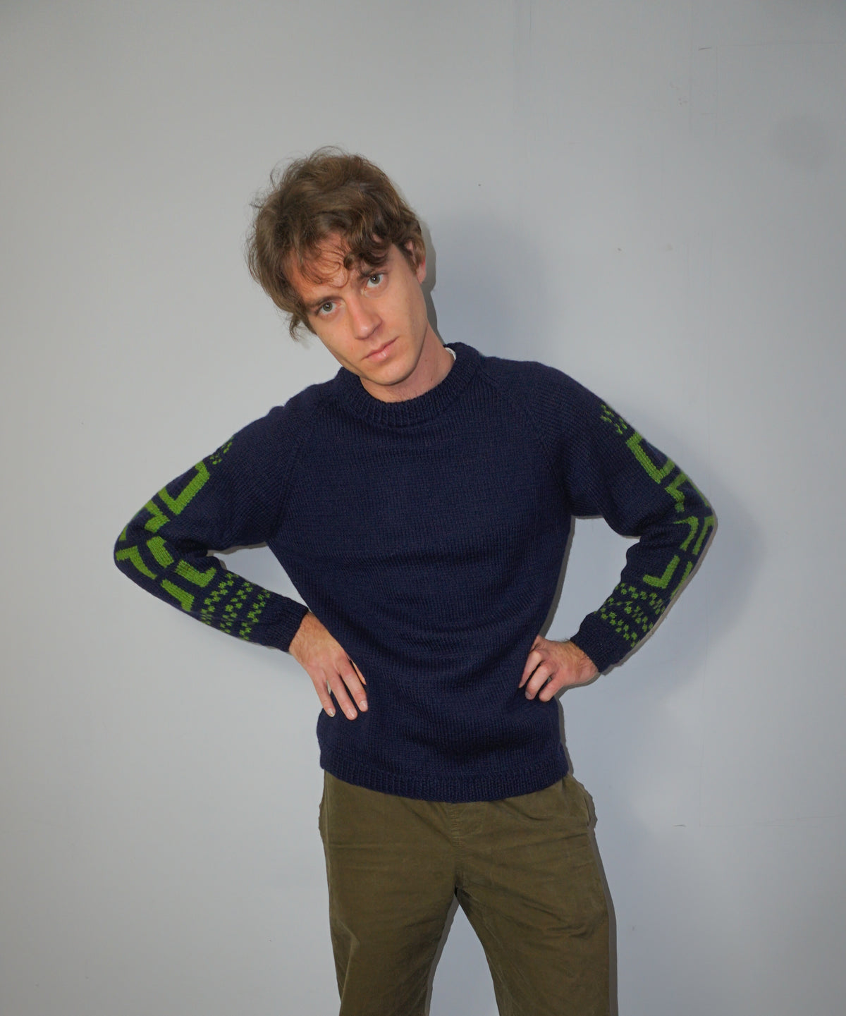 A man wearing a navy blue knit sweater with green geometric patterns on the sleeves, paired with olive green pants, posing against a plain light gray wall.