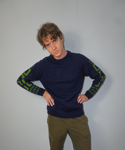 A man wearing a navy blue knit sweater with green geometric patterns on the sleeves, paired with olive green pants, posing against a plain light gray wall.
