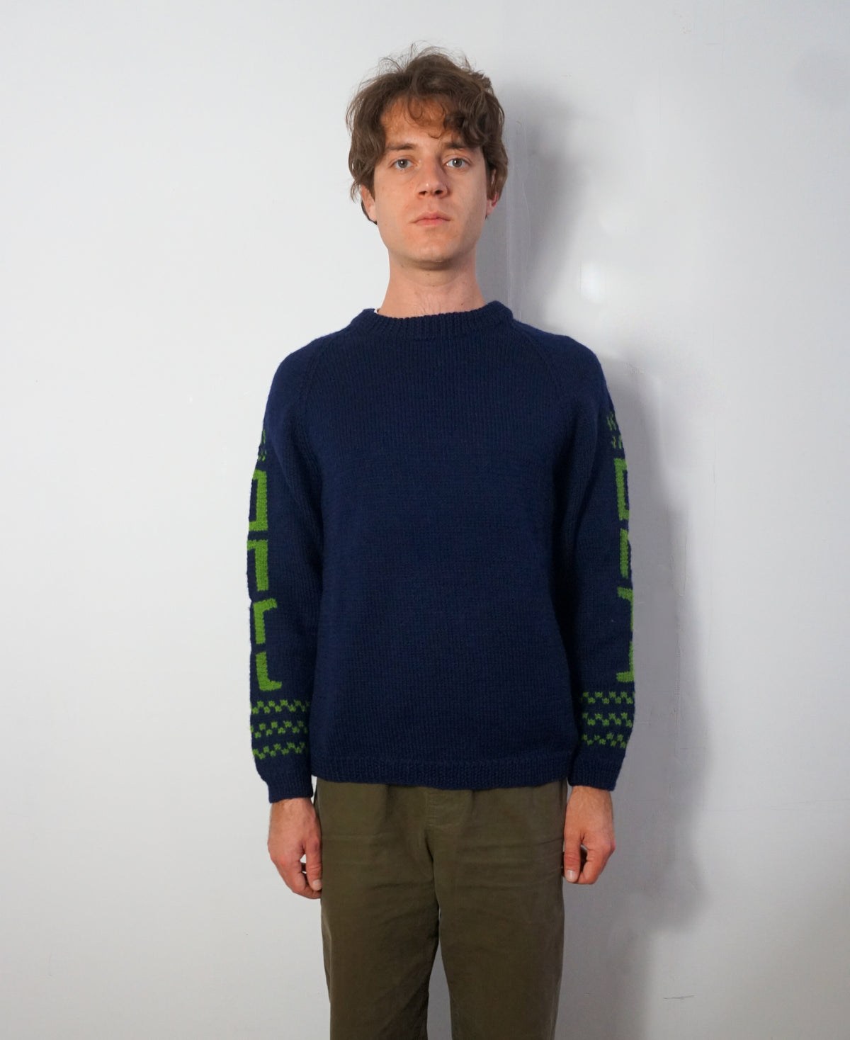 A man wearing a navy blue knit sweater with green geometric patterns on the sleeves, paired with olive green pants, standing against a plain light gray wall.