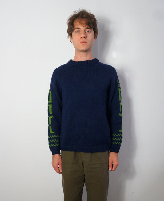 A man wearing a navy blue knit sweater with green geometric patterns on the sleeves, paired with olive green pants, standing against a plain light gray wall.