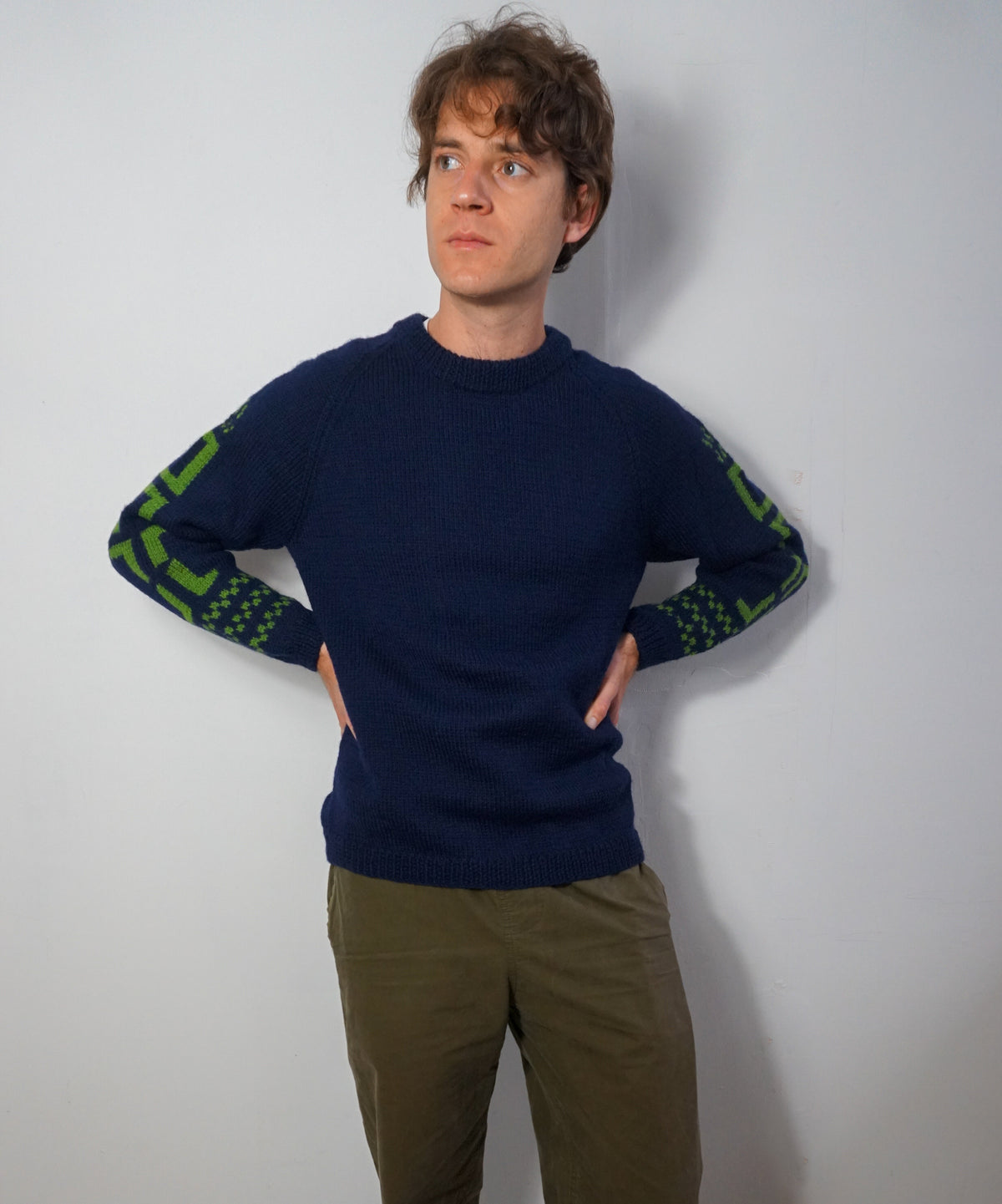 A man wearing a navy blue crewneck sweater with green patterned knit detailing on the sleeves, paired with olive green pants, standing against a plain light gray wall.