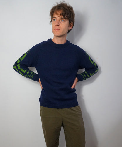 A man wearing a navy blue crewneck sweater with green patterned knit detailing on the sleeves, paired with olive green pants, standing against a plain light gray wall.