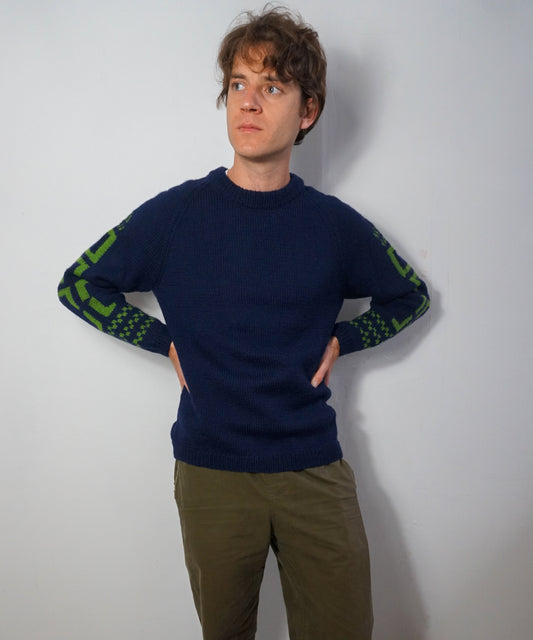 A man wearing a navy blue crewneck sweater with green patterned knit detailing on the sleeves, paired with olive green pants, standing against a plain light gray wall.