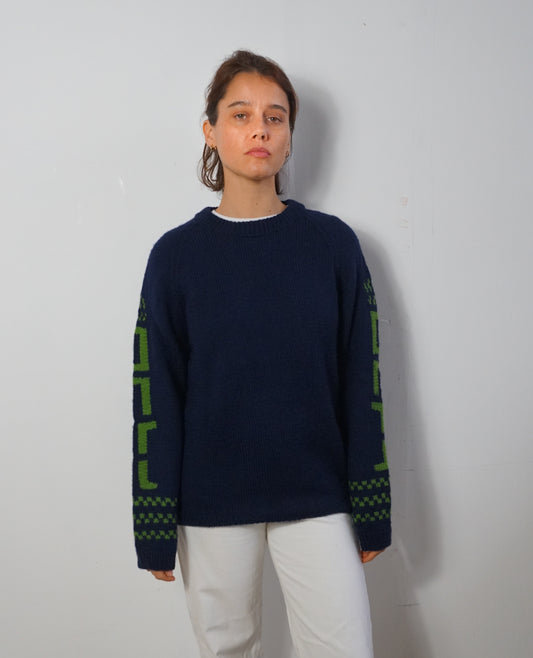 A woman wearing a navy blue knit sweater with green geometric patterns on the sleeves, paired with white pants, standing against a plain light gray wall.