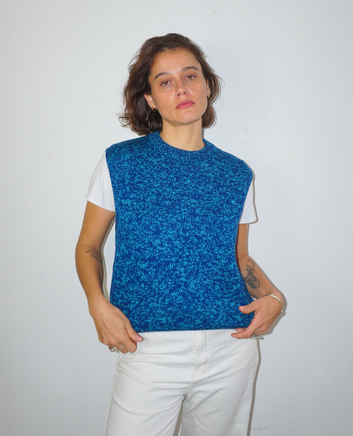 A woman modeling a sleeveless, textured blue knit vest over a white t-shirt and white pants, standing against a plain white wall.