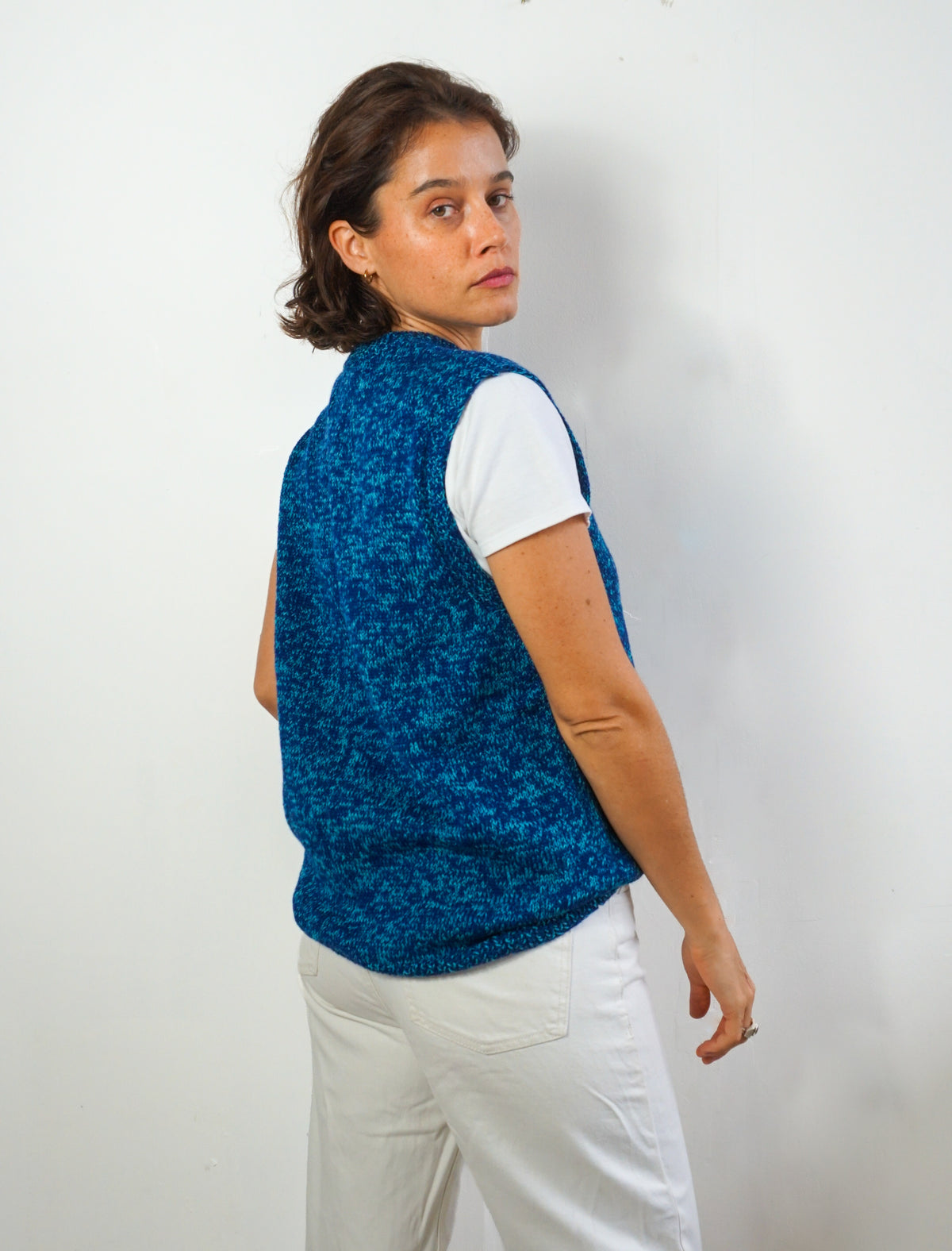 A woman wearing a blue textured knit sleeveless vest over a white t-shirt and white pants, standing against a plain white wall.