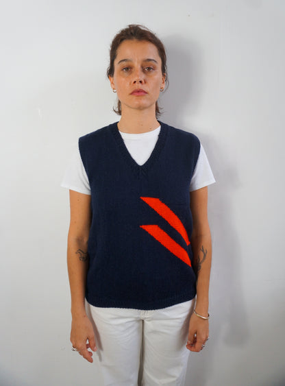 A woman stands against a plain light gray wall wearing a navy blue knit sweater vest with a bold red diagonal stripe graphic on the front, layered over a white t-shirt and white pants.