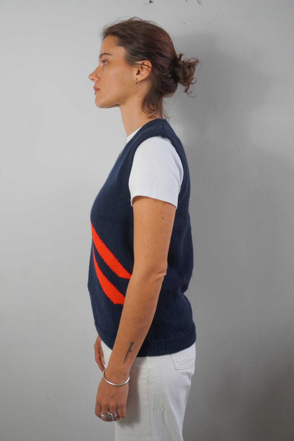 A woman shown in profile wearing a navy blue knit sweater vest with two diagonal orange stripes, layered over a white t-shirt and white pants, standing against a plain light gray wall.