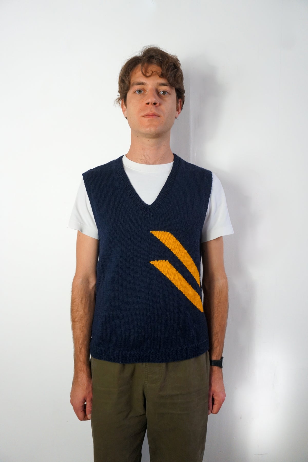 A man wearing a navy blue knit sweater vest with a yellow chevron pattern over a white t-shirt and olive green pants, standing against a plain white wall.
