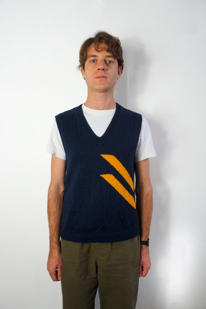 A man wearing a navy blue knit sweater vest with a yellow chevron pattern over a white t-shirt and olive green pants, standing against a plain white wall.