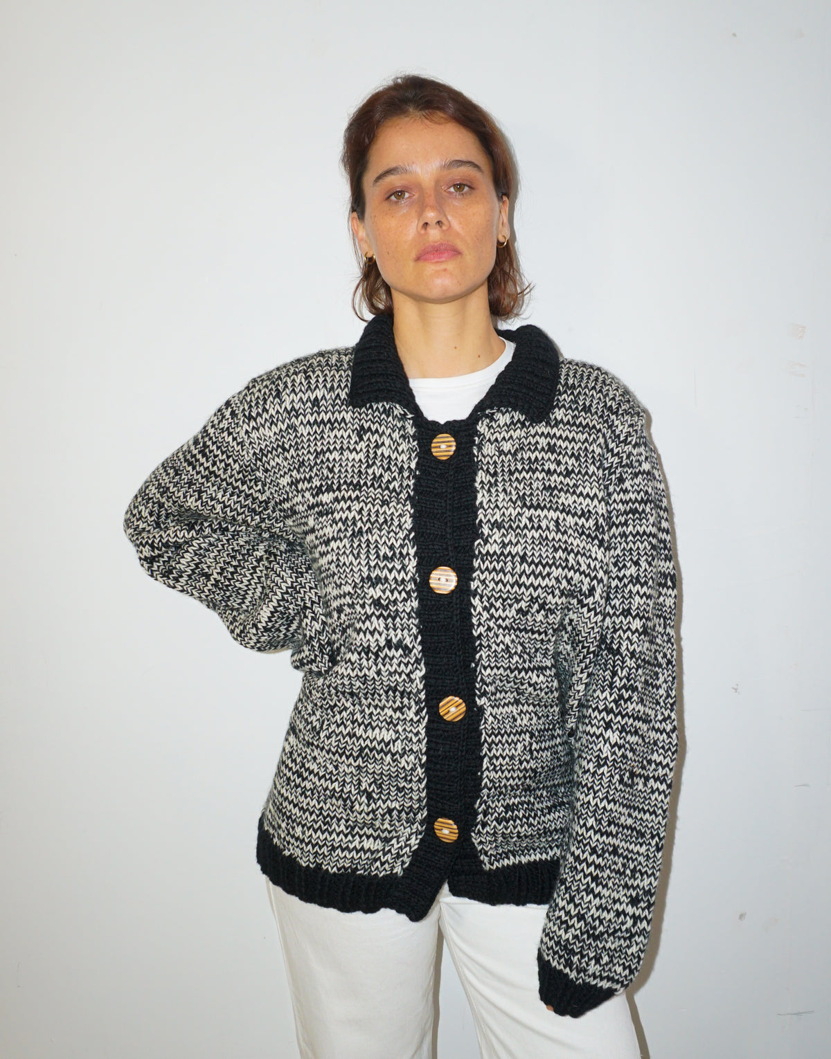 A woman wearing a black and white marled knit cardigan with black trim and gold buttons, paired with white pants, standing against a plain white wall.