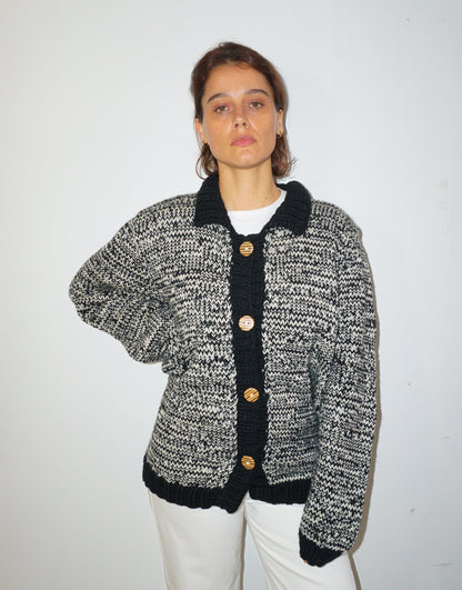 A woman wearing a black and white marled knit cardigan with black trim and gold buttons, paired with white pants, standing against a plain white wall.