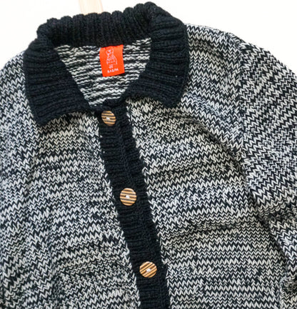 A close-up of a knit cardigan sweater with a black ribbed collar and button placket, featuring a black, gray, and white wavy patterned body, and wooden buttons.
