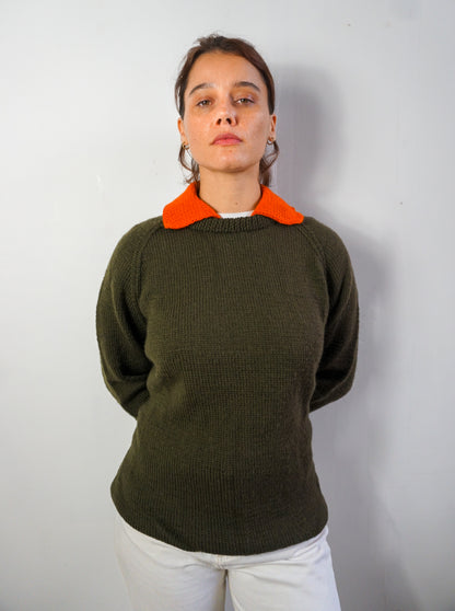 A woman wearing a dark olive green knit sweater with a contrasting bright orange collar, paired with white pants, standing against a plain light gray wall.