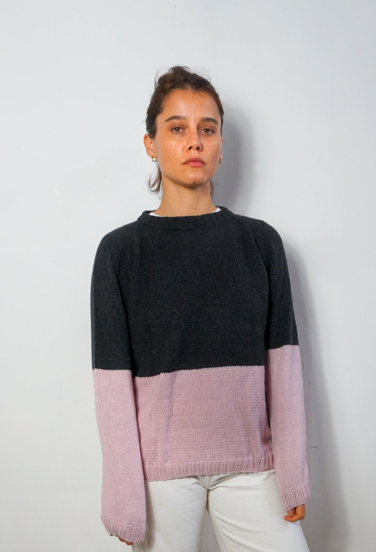 A woman wearing a color-block knit sweater with a dark charcoal upper half and light pink lower sleeves and hem, paired with white pants, standing against a plain light gray wall.