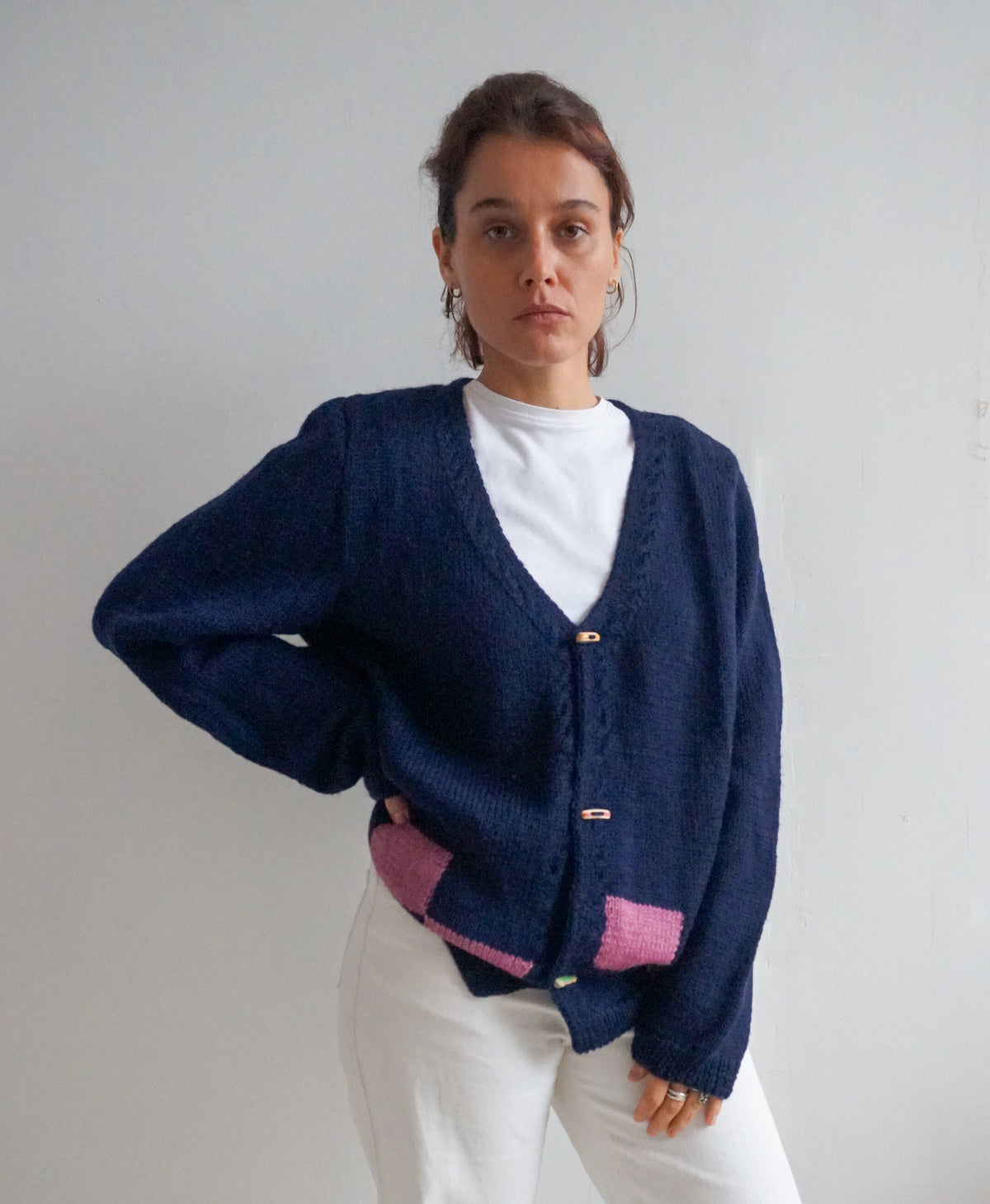 A woman wearing a navy blue knit cardigan with pink color-blocked details at the hem and cuffs, layered over a white t-shirt and white pants, standing against a plain light gray wall.
