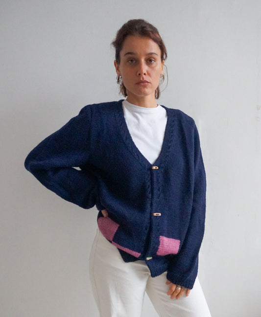 A woman wearing a navy blue knit cardigan with pink color-blocked details at the hem and cuffs, layered over a white t-shirt and white pants, standing against a plain light gray wall.