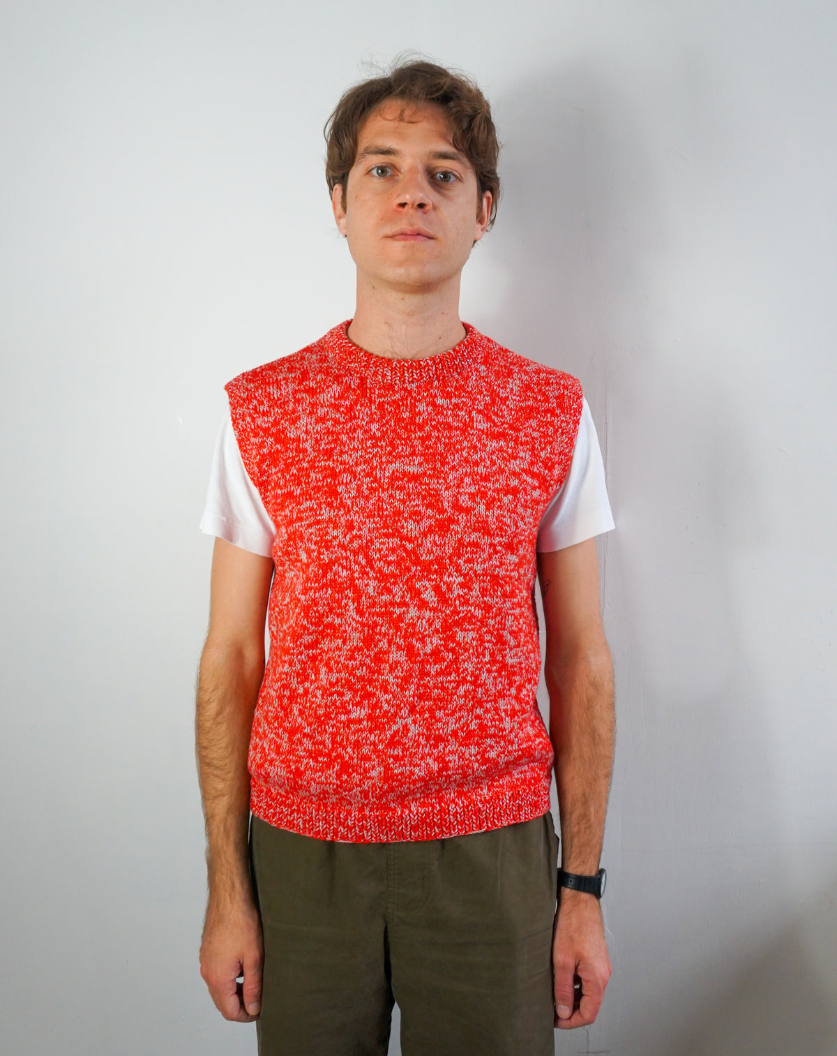 A man wearing a bright red and white marled knit sweater vest over a white t-shirt and olive green pants, standing against a plain light gray wall.