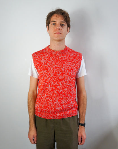 A man wearing a bright red and white marled knit sweater vest over a white t-shirt and olive green pants, standing against a plain light gray wall.