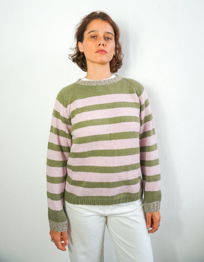 A woman wearing a green and pink striped knit jumper, paired with white pants, standing against a plain white wall.