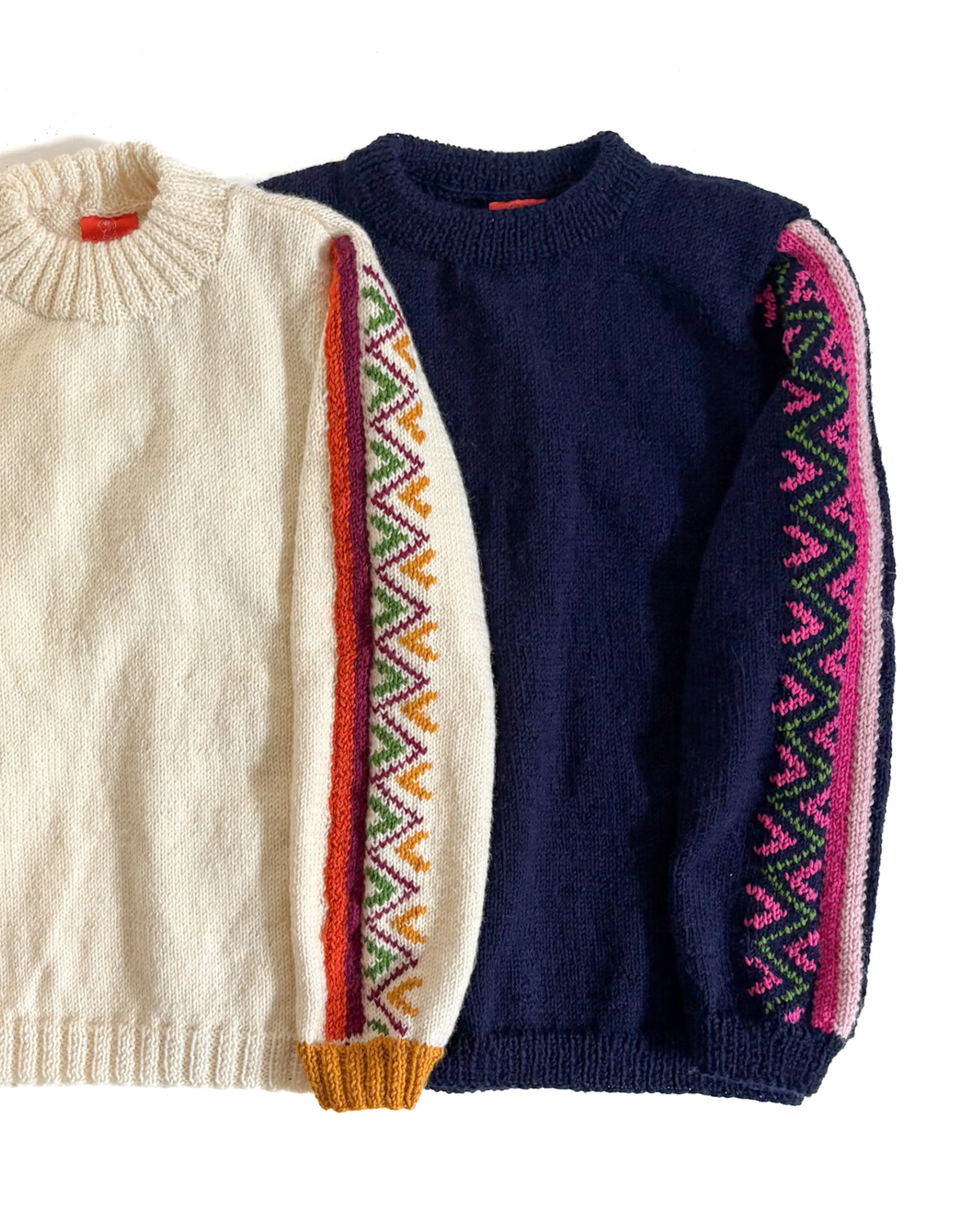 Two knitted crewneck sweaters laid flat on a white background. The left sweater is cream and the other one is navy with a multicolor geometric pattern down the sleeve.