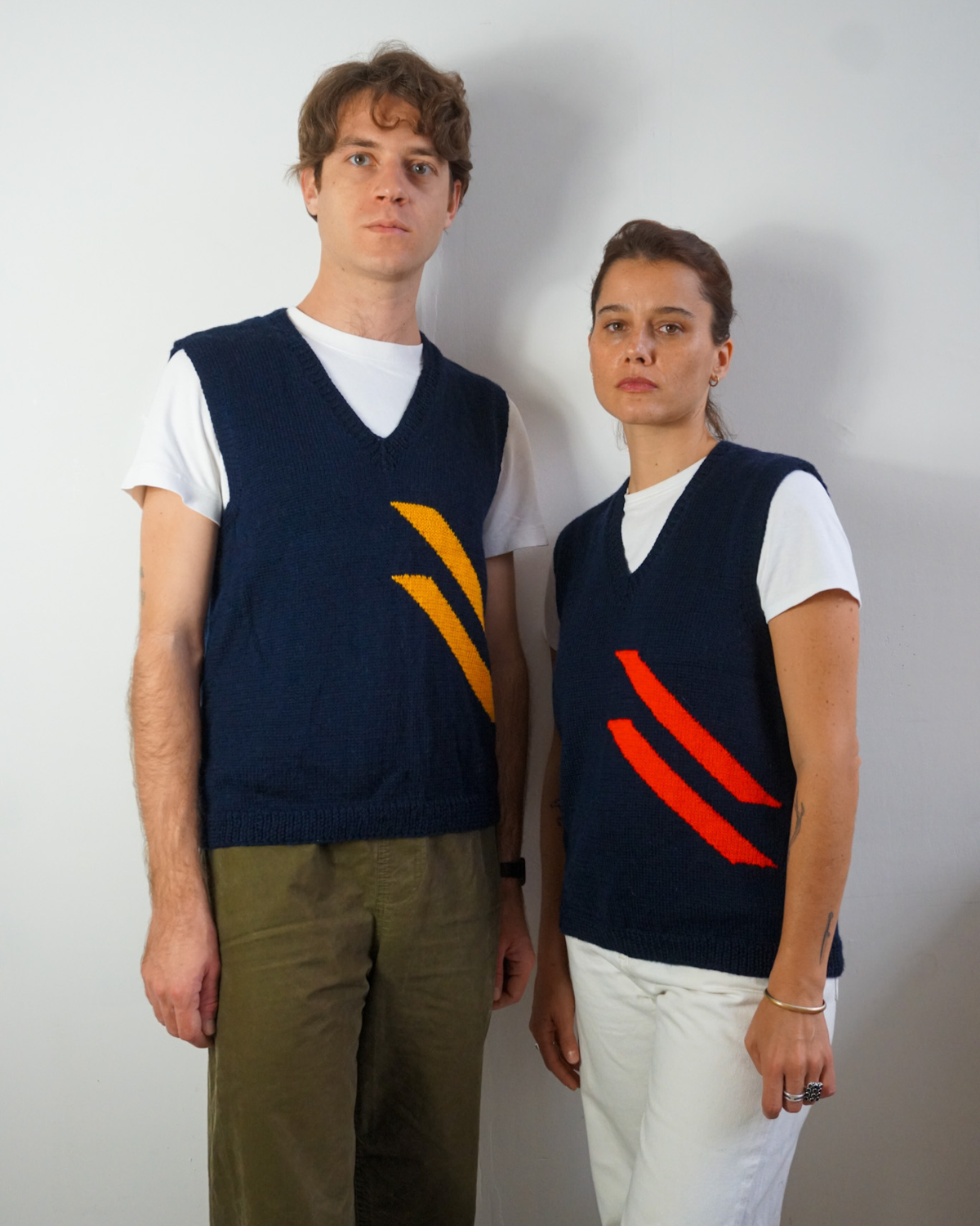 Two models wearing matching navy blue V-neck knit sweater vests with diagonal stripe patterns in yellow and red, layered over white t-shirts and paired with olive green and white pants, standing against a plain light gray wall.