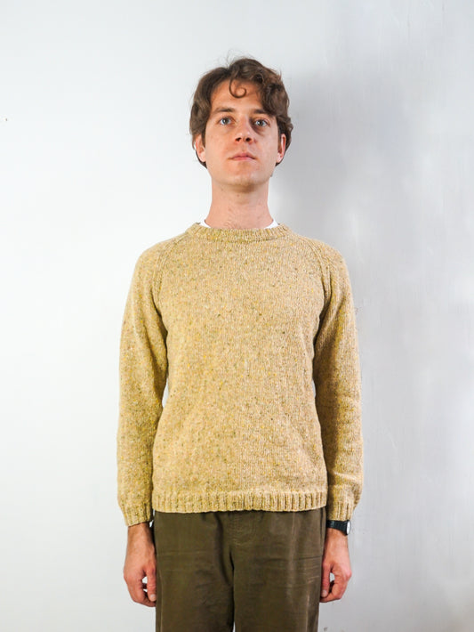 A man wearing a textured, hand-knit crewneck sweater in a warm mustard yellow-beige color, paired with olive green trousers, standing against a plain white studio background.